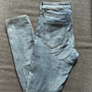 Ljusblå jeans i storlek 38 - Säljer ett par ljusblå jeans i klassisk femficksmodell. Jeansen har normal passform och raka ben, med snygga slitningar och detaljerade sömmar. Perfekta för dig som gillar en enkel och stilren look. Stängs med dragkedja och knapp.