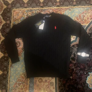 Svart kabelstickad tröja Polo Ralph Lauren - Svart kabelstickad tröja från Polo Ralph Lauren med klassisk röd broderad logga på bröstet. Tröjan har rund halsringning och långa ärmar. Tillverkad i mjuk bomull, perfekt för lager-på-lager under höst och vinter.