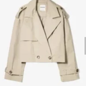 Beige croppad trenchcoat från Bershka - Snygg croppad trenchcoat från Bershka i beige färg. Jackan har bred krage, dubbla knapprader och detaljer med knappar vid axlar och ärmslut. Tillverkad i bomullsmaterial och har en loose passform som ger en cool vibe.