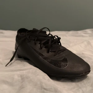 Svarta Nike fotbollsskor strl 43 - Säljer ett par svarta Nike fotbollsskor med snörning och rund tå. Skorna har en syntetisk ovandel och platt sula med dobbar för bra grepp på planen. Snygg, stilren design med diskret Nike-logga på sidan. Perfekt för dig som vill ha grymma skor till matchen.