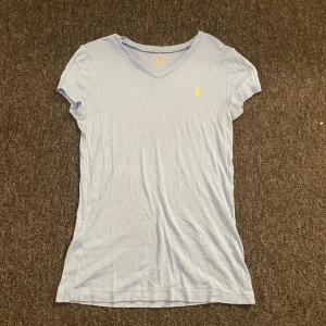 Ljusblå t-shirt från Polo Ralph Lauren - Snygg ljusblå t-shirt från Polo Ralph Lauren med klassisk gul broderad logga på bröstet. T-shirten har v-ringad hals och korta ärmar, tillverkad i mjuk bomull som känns skön mot huden. Perfekt för en chill och stilren look. Passar någon som har XS/S.