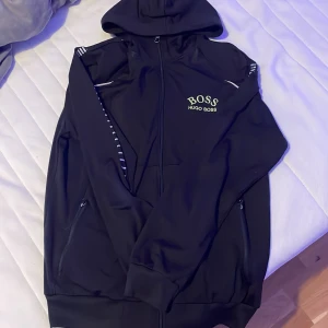 Svart Hugo Boss hoodie med dragkedja - Svart hoodie från Hugo Boss med broderad logga på bröstet i vitt. Tröjan har hel dragkedja, huva och diskreta vita detaljer på axlarna. Tillverkad i mjukt syntetmaterial och har långa ärmar samt fickor med dragkedja.