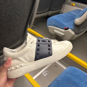 Valentino Open sneakers med nitar - Snygga vita Valentino Open sneakers i skinn med marin blå band och ikoniska pyramidformade nitar. Klassisk rund tå och platt sula. Perfekta för dig som vill ha en lyxig och edgy look. Kommer med originalkartong.