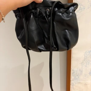 Svart handväska med snörning - Cool svart handväska i mjukt skinnliknande material med snygg snörning upptill. Väskan har en puffig, avslappnad form och långa remmar som ger en trendig vibe. Perfekt för dig som vill ha en stilren men ändå unik accessoar.