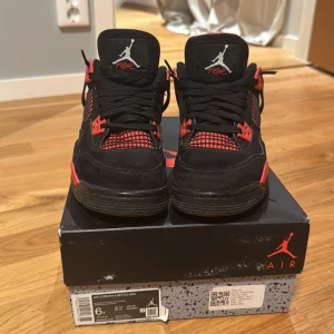 Nike Air Jordan 4 Retro svart/röd - Nike Air Jordan 4 Retro sneakers i svart och rött med ikonisk Jumpman-logga på hälen och plösen. Skorna har meshpaneler, snörning och detaljer i syntet och mocka. Snygg kontrast mellan den svarta ovandelen och den röda sulan samt insidan.