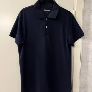 Mörkblå pikétröja från Kappahl S - Snygg mörkblå pikétröja från Kappahl i storlek S. Klassisk modell med krage, två knappar och korta ärmar. Den har aldrig använts och är i helt nytt skick. Passar perfekt till när man vill vara lite extra uppklädd. Hör av er om ni har frågor!