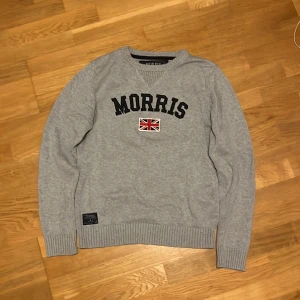 Grå stickad tröja från Morris - Snygg grå stickad tröja från Morris med broderad logga och brittisk flagga på bröstet. Tröjan har rund halsringning, ribbade muddar och en klassisk passform. Perfekt för kallare väder och en skön look! Mycket fint skick och storlek Medium, hör av dig vid några frågor, priset kan diskuteras!