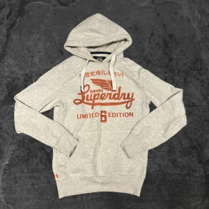 Grå hoodie från Superdry - Snygg ljusgrå hoodie från Superdry med röd logga och japanska tecken framtill. Tröjan har huva med snörning, magficka och ribbade muddar. Perfekt för dig som gillar streetstyle och vill ha något bekvämt och stilrent.