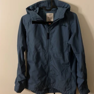 Blå vindjacka från Lundhags med huva - Snygg blå vindjacka från Lundhags i juniorstorlek 158/164. Jackan har huva, dragkedja framtill och diskret broderad logga på bröstet. Perfekt för blåsiga dagar och outdoor-äventyr. Materialet är slitstarkt och känns lätt på kroppen. Lite små fläckar där fram men går säkert bort i tvätt.