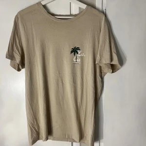 Beige t-shirt från Jack & Jones - Snygg beige t-shirt från Jack & Jones med liten palmtryck och texten 'L.A.' på bröstet. Klassisk rund hals och korta ärmar. Perfekt basic-plagg med clean look och soft känsla. Tillverkad i mjuk bomull för skön komfort.