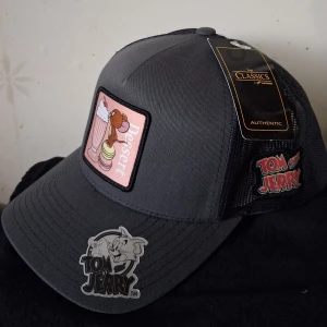 Tom & Jerry grå truckerkeps - Snygg grå truckerkeps med Tom & Jerry-motiv och mesh på sidorna. Framtill finns en patch med Jerry och milkshake i rosa toner samt broderad Tom & Jerry-logga. Justerbar snapback och böjd skärm. Ny,oanvänd.