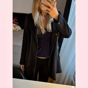 Mörkgrå ull kappa med bälte från Vero Moda - Snygg mörkgrå kappa från Vero Moda med bred krage, långa ärmar och bälte i midjan. Jackan har två dragkedjeförsedda fickor och stängs med både knappar och bälte. Perfekt för dig som gillar stilren och modern look. Kappan har ej använts och har prislappen på. Jackans material är gjord av ull. Nypris: 1000kr