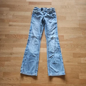 Bootcut ljusblå jeans med slitningar från Gina Tricot. - Snygga ljusblå bootcut jeans med slitningar på knäna och coola fickor med lock och knapp bak. Jeansen har klassisk femficksdesign och är tillverkade i jeansmaterial. Perfekt för dig som gillar en avslappnad och trendig stil.