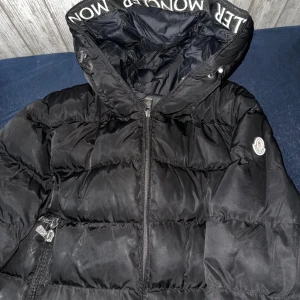 Svart Moncler pufferjacka med huva - Svart pufferjacka från Moncler med stor huva och logotyp på ärmen. Jackan har dragkedja framtill, dubbla dragkedjor och Moncler-tryck på huvkanten. Fodrad och riktigt fluffig, perfekt för kalla dagar. Klassisk design med snygga detaljer och innerfoder med serietidningsmönster.
