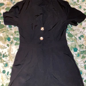 Svart kortklänning från Suffle - Svart kortklänning från Suffle med två snygga silvriga knappar framtill. Klänningen har korta ärmar och markerad midja, vilket ger en clean och stilren look. Tillverkad i ett mjukt och lätt material som känns bekvämt mot huden. Det är inte en klänning typ men det är en klänning fast med shorts. 