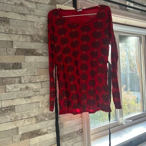 Röd långärmad  med äppelmönster från Me&I - Säljer en röd långärmad  från Me&I i storlek M med svart äppelmönster och rund halsringning. Klänningen har långa ärmar och ett  knytband i midjan som ger en snygg siluett. Materialet känns mjukt och stretchigt, perfekt för dig som gillar färg och mönster.