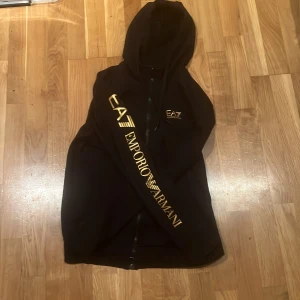 Svart EA7 Emporio Armani hoodie S - Svart hoodie från EA7 Emporio Armani med guldfärgat tryck på bröstet och längs ena ärmen. Tröjan har dragkedja framtill, huva med snörning och ribbade muddar. Perfekt för dig som gillar streetwear och vill sticka ut med exklusiva detaljer.