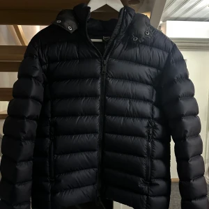 Mörkblå pufferjacka från Colmar - Säljer en mörkblå pufferjacka från Colmar med avtagbar huva och dragkedja framtill. Jackan har en quiltad look med horisontella sömmar och två fickor med dragkedja. Perfekt för kyliga dagar. Köpt för 4500kr har kvitto! Använd endast en säsong så i väldigt gott skick!