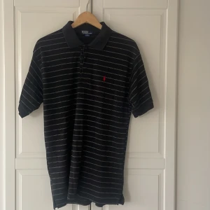 Svart randig piké från Polo Ralph Lauren - Svart pikétröja från Polo Ralph Lauren i storlek XL. Tröjan har tunna vita ränder, klassisk krage och knappar framtill. Materialet är 100% bomull och på bröstet finns den ikoniska röda broderade loggan. Perfekt för en chill och stilren look.