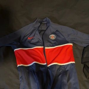 PSG Nike träningsjacka marinblå/röd S - Säljer en snygg PSG träningsjacka från Nike i marinblått och rött med vit detalj. Jackan har dragkedja, lång ärm och är gjord i Dri-Fit polyester. Perfekt för fotboll eller chill. PSG-logga och Nike Swoosh på bröstet.