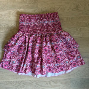 Rosa mönstrad kortkjol med smock - Supersöt kortkjol i rosa med färgglatt paisleymönster och bred smock i midjan. Kjolen har en volangig och luftig form som ger en skön och avslappnad vibe. Perfekt för dig som gillar färg och mönster! Materialet känns lätt och passar bra till varmare dagar.