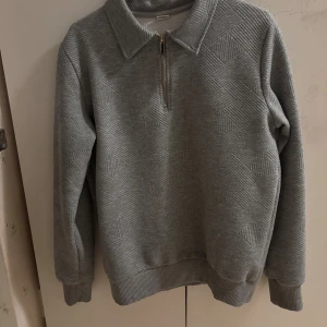 Grå halvzip tröja med strukturmönster - Grå långärmad tröja med halv dragkedja och krage. Tröjan har ett snyggt strukturmönster i tyget och ribbade muddar vid ärmslut och nederkant. Perfekt för lager-på-lager och chill dagar.