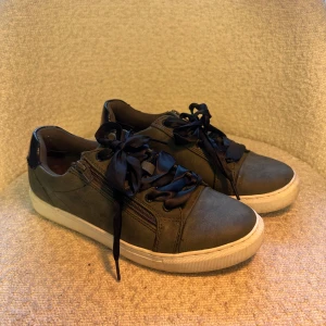 Mörkgröna sneakers med satinband storlek 38 - Snygga mörkgröna sneakers med platt vit sula och rund tå. Skorna har breda satinband som snörning och dragkedja på sidan för extra detalj. Materialet är mocka-liknande och insidan är mjukt textilfoder. Perfekt för dig som gillar stilrena och coola detaljer.