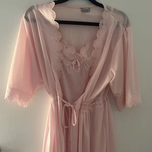 Ljusrosa sov omlottklänning från Yunyny - Supersöt ljusrosa omlottklänning från Yunyny med spetsdetaljer längs kanterna och ärmarna. Klänningen har knyt i midjan och v-ringad hals med fina broderade spetsmönster. Perfekt för dig som gillar romantisk och feminin stil.