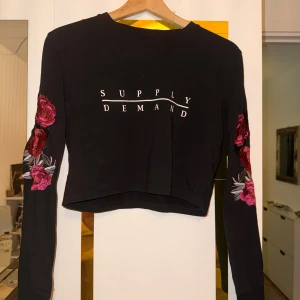 Supply & Demand croppad svart tröja - Svart croppad långärmad tröja från Supply & Demand med vit text fram och bak. Snygga broderade röda och rosa blommor på ärmarna ger en unik vibe. Perfekt för dig som gillar streetstyle och detaljer. Mjuk bomullskänsla och normal passform.