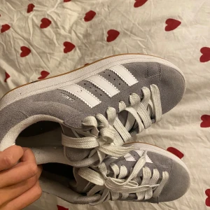 Grå Adidas Campus sneakers - Säljer ett par grå Adidas Campus sneakers med klassiska vita ränder på sidorna och vit snörning. Skorna har en låg profil, rund tå och platt sula i gummi. Ovandelen är i mjuk mocka och insidan är fodrad med textil. Perfekt för dig som gillar retrostil och streetwear. Köpte dem ganska nyligen och är bara använda två gånger. 