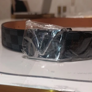 Louis Vuitton rutigt svart bälte - Snyggt Louis Vuitton bälte med klassiskt LV-spänne i silverfärg. Bältet har ett rutigt mönster i svart och grått, och är tillverkat i skinn med brunt insida. Perfekt accessoar för att lyfta din outfit med exklusiv känsla och stil.