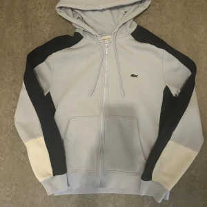 Ljusgrå hoodie från Lacoste med dragkedja - Snygg ljusgrå hoodie från Lacoste med klassisk krokodillogga på bröstet. Tröjan har svarta och vita paneler på ärmarna, huva med snörning och hel dragkedja framtill. Perfekt för chill eller streetwear. Materialet känns mjukt och är troligen bomull eller polyester.