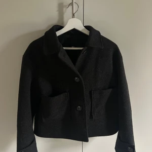 Svart kappa - Stilren svart kappa i ullmaterial. Jackan har en rak, croppad passform med stora framfickor, klassisk krage och knäppning med stora knappar framtill. Perfekt för dig som gillar minimalistisk och clean stil.