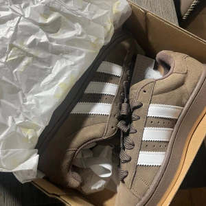 Adidas Campus sneakers brun/vit - Säljer ett par Adidas Campus sneakers i brun mocka med klassiska vita ränder på sidorna. Skorna har rund tå, platt gummisula i beige och snörning med breda bruna skosnören. Perfekt för dig som gillar retrostil och snygga detaljer.