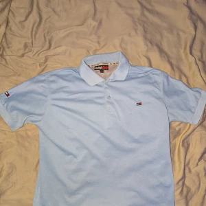 Ljusblå pikétröja Tommy Hilfiger  - Säljer en ljusblå pikétröja från Tommy Hilfiger i storlek S. Klassisk krage, korta ärmar och knäppning framtill. Ikoniska Hilfiger-loggor på bröstet och ärmen. Perfekt för en clean och stilren look. 
