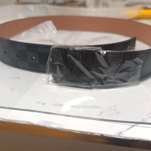 Svart rutig Louis Vuitton skärp - Säljer ett stilrent Louis Vuitton bälte med klassiskt rutigt mönster i svart och grått. Bältet har en snygg LV-logga på spännet och är tillverkat i skinn med beige insida. Perfekt för dig som vill ha en lyxig detalj till outfiten.