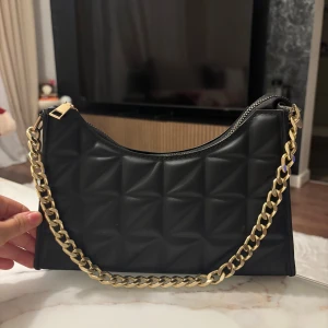 Svart  axelväska med guldkedja - Snygg svart axelväska med quiltad design och en chunky guldfärgad kedja som axelrem. Väskan har dragkedja upptill och är tillverkad i skinnimitation. 