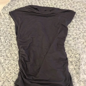 Mörkgrå topp från Gina Tricot - Mörkgrå topp från Gina Tricot i mjuk bomull. Modellen är ärmlös med lätt rynkade detaljer på sidan som ger en snygg passform. Enkel och stilren design som är lätt att matcha med accessoarer.