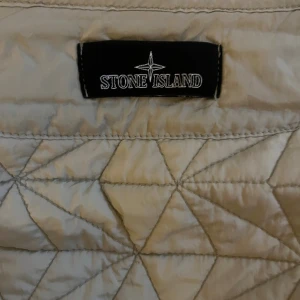 Quiltad beige väst från Stone Island - Tar högsta bud efter någon dag. Snygg quiltad väst från Stone Island i beige färg med geometriskt mönster. Västen har dragkedja framtill och tryckknappar vid kragen. Perfekt att slänga över en hoodie för extra stilpoäng. Materialet är polyester och designen är riktigt clean.