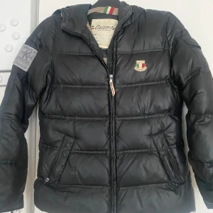 Svart pufferjacka från Dolomite S - Svart pufferjacka från Dolomite med broderad logga på bröstet och stor brodyr på ryggen. Jackan har huva, dragkedja framtill och två snedställda fickor. Materialet är glansigt och känns sportigt, perfekt för kalla dagar. Snygga detaljer med italienska flaggan på dragkedjan.