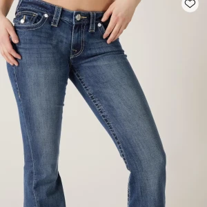 True Religion blå bootcut jeans - Snygga blå jeans från True Religion med bootcut passform och klassisk gul kontrastsöm. Jeansen har låg midja, fem fickor och ikonisk broderad bakficka med knapp. Materialet är slitstarkt denim i bomull. Perfekt för dig som gillar en cool och avslappnad stil. Nypris 1300