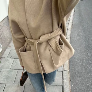HM Beige omlott kappa med bälte - Säljer en beige omlott kappa med bälte i midjan och stora framfickor. Jackan har en clean look utan krage och är tillverkad i ett mjukt, ulligt material. Perfekt för dig som gillar minimalistisk stil och vill ha en snygg jacka till hösten.