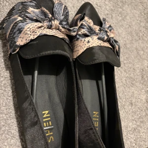 Svarta ballerinaskor med rosett från SHEIN - Snygga svarta ballerinaskor från SHEIN med spetsig tå och en stor rosett framtill i beige och svart mönster. Skorna har en platt sula och är tillverkade i syntetmaterial med en satinliknande finish. Perfekta för dig som vill ha något extra till outfiten.