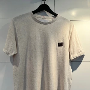Beige t-shirt från Les Deux - Snygg beige t-shirt från Les Deux med rund hals och diskret broderad patch med romerska siffror II på bröstet. Tillverkad i mjuk bomull, perfekt för en clean och stilren look.