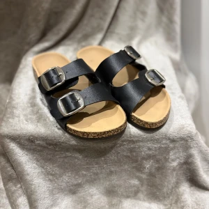Svarta sandaler med dubbla spännen - Svarta sandaler med två breda remmar och stora silverspännen. Fotbädden är ljusbeige och sulan är i kork-look med svart undersida. Perfekta för varma dagar och enkel att matcha med allt. Klassisk och stilren design.