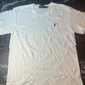 Vit t-shirt från Polo Ralph Lauren - Vit t-shirt från Polo Ralph Lauren med klassisk rund hals och korta ärmar. På bröstet finns den ikoniska broderade loggan i svart. T-shirten är gjord i mjuk bomull och har en clean, stilren look som funkar till allt.
