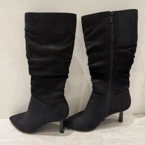 Svarta stövlar med spetsig tå och klack - Snygga svarta stövlar i mocka-look med spetsig tå och smal klack. Stövlarna har dragkedja på insidan och en stilren, slouchy design som ger en trendig vibe. Perfekta för att lyfta vilken outfit som helst.