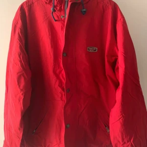 Röd vindjacka från Chinook Sport - Röd vindjacka från Chinook Sport med huva och blå snörning. Jackan har dragkedja och tryckknappar framtill, samt två fickor med dragkedja. Lite gulnad på insidan kragen, men inget som syns när man har den på sig. Går säkert att få bort med lite omsorg. 