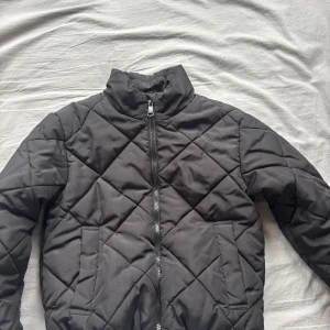 Svart quiltad pufferjacka Samsøe Samsøe - Svart pufferjacka från Samsøe Samsøe med quiltat mönster och hög krage. Jackan har dragkedja framtill, två fickor och lång ärm. Perfekt för kyliga dagar och ger en clean, modern look.