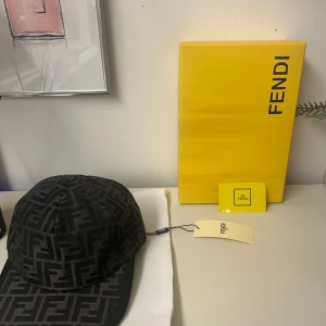 Svart Fendi keps med FF-mönster - Snygg svart keps från Fendi med det klassiska FF-logomönstret över hela kepsen. Kepsen har böjd skärm och justerbar rem baktill med Fendi Roma-detalj i metall. Materialet är bomull och polyester, vilket gör den både bekväm och stilren.
